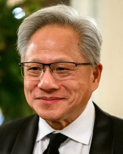 Jensen Huang smiling, with grey hair and glasses, in a dark suit