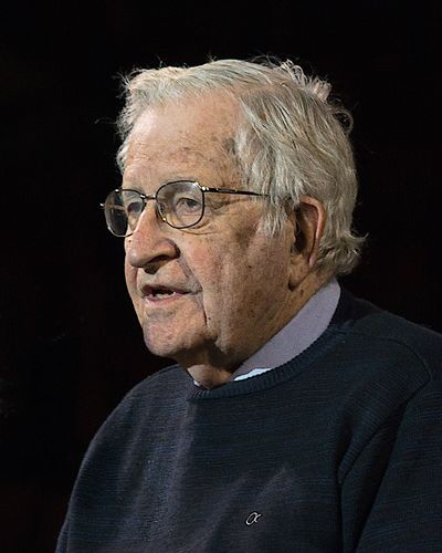Noam Chomsky, an older man with white hair and glasses, in a dark sweater, speaking